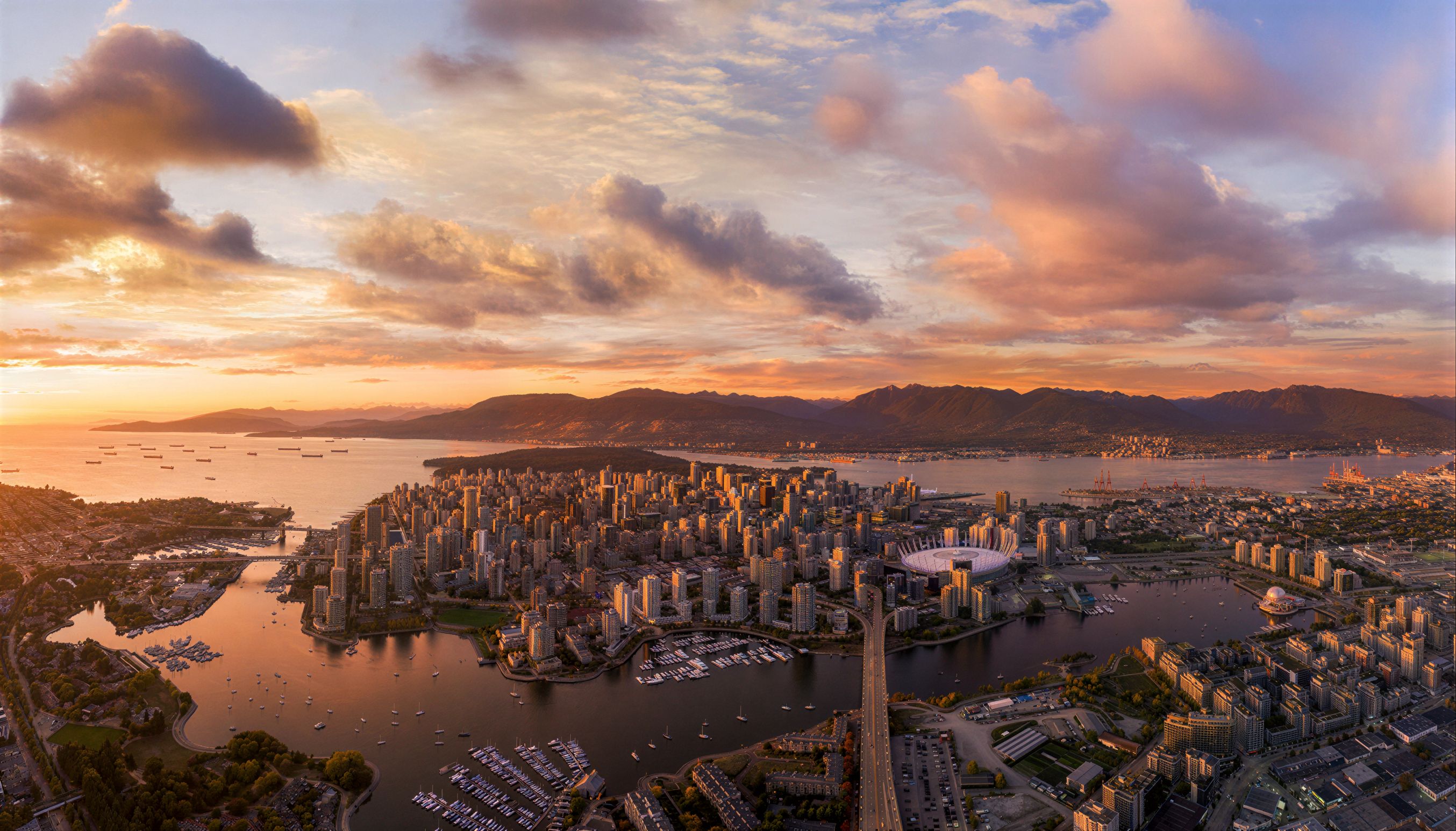 Aerial view of False Creek at sunset, with Concord Pacific Place's waterfront towers along the seawall, BC Place stadium at centre, and the North Shore mountains beyond